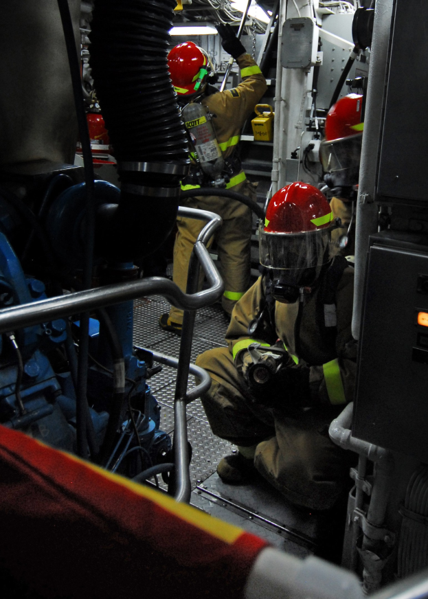 Feuerwehrleute in Helmen und Schutzausrüstung arbeiten an einem Feuerwehraufsatz mit Rohren und einer Lampe im Hintergrund.