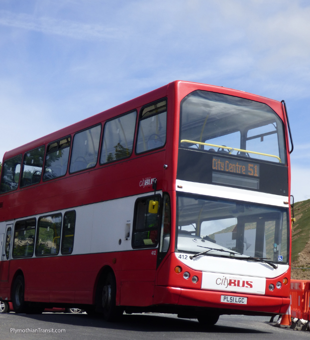 Ein roter Doppeldeckerbus mit der Aufschrift "Stadtbus" fährt auf der Straße, daneben steht ein Verkehrskegel und im Hintergrund ist ein Hügel zu sehen.