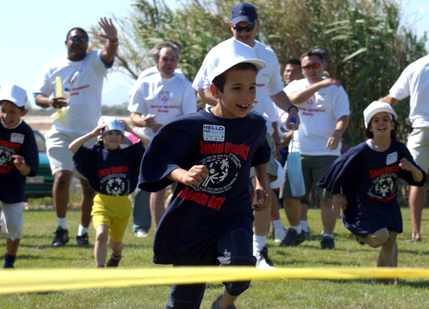 Gruppe von Kindern, die auf einem Rasenfeld einen Lauf Wettbewerb absolvieren, mit einem gelben Band im Vordergrund, einige tragen Kappen und Schutzbrillen, andere halten Gegenstände, Bäume und ein klarer blauer Himmel im Hintergrund.