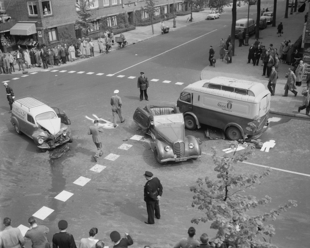 Eine Schwarz-Weißt-Aufnahme eines Autounfalls an einer Straßenkreuzung, mit mehreren Fahrzeugen, darunter ein Lieferwagen, einer Gruppe von Menschen drumherum und im Hintergrund Gebäude, Bäume, Masten und Texttafeln.