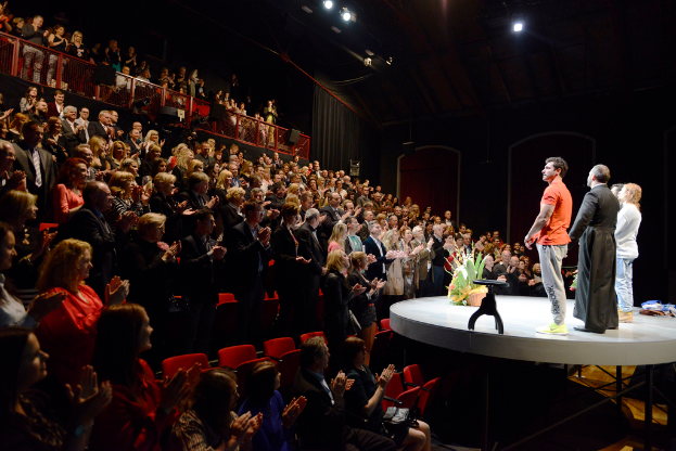 Eine Gruppe von Menschen, die auf einem Boden stehen, einige sitzen im Vordergrund auf Stühlen, ein Blumenkorb und ein Hocker auf der Bühne rechts und Lautsprecher, ein Metallgitter und Lichter im Hintergrund.