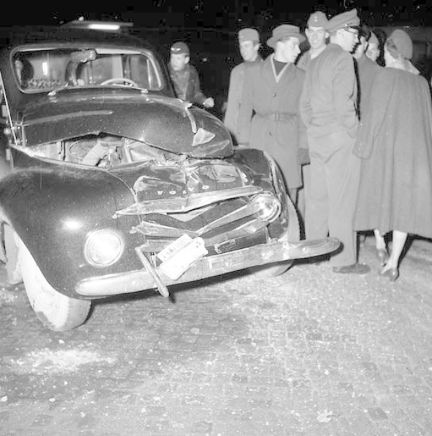 Schwarz-weißes Bild eines schwer beschädigten Autos am Straßenrand mit eingedellter Motorhaube und zersplitterten Fenstern, umgeben von Menschen, die die Szene begutachten.