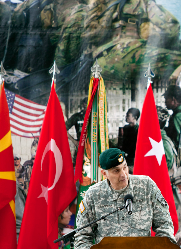 Ein Soldat steht an einem Podium mit einem Mikrofon, mit verschiedenen Flaggen und einem Plakat von Militärpersonal im Hintergrund.