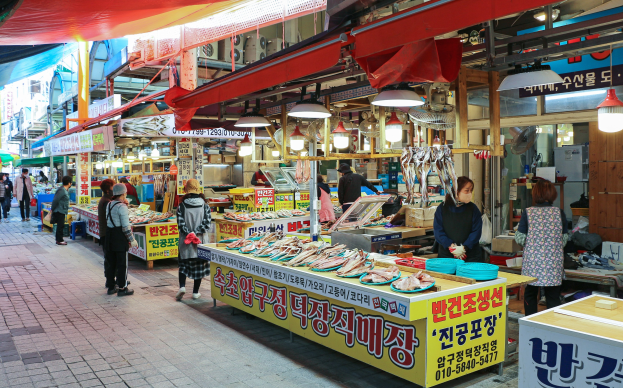 Ein belebter Straßenmarkt in Seoul, Südkorea mit Menschen, die gehen, Ständen mit verschiedenen Gegenständen und Gebäuden im Hintergrund bei klarem blauem Himmel.