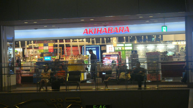 Ein Geschäft mit zwei Bänken davor, auf denen jeweils einige Personen sitzen, einem Schild darüber und einer eingezäunten Wand unten.