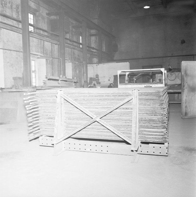 Ein Schwarz-Weiß-Foto einer Eisengießerei mit Metallobjekten auf dem Boden, einer Tür auf der rechten Seite, einer Wand mit Fenstern im Hintergrund und Lampen oben.