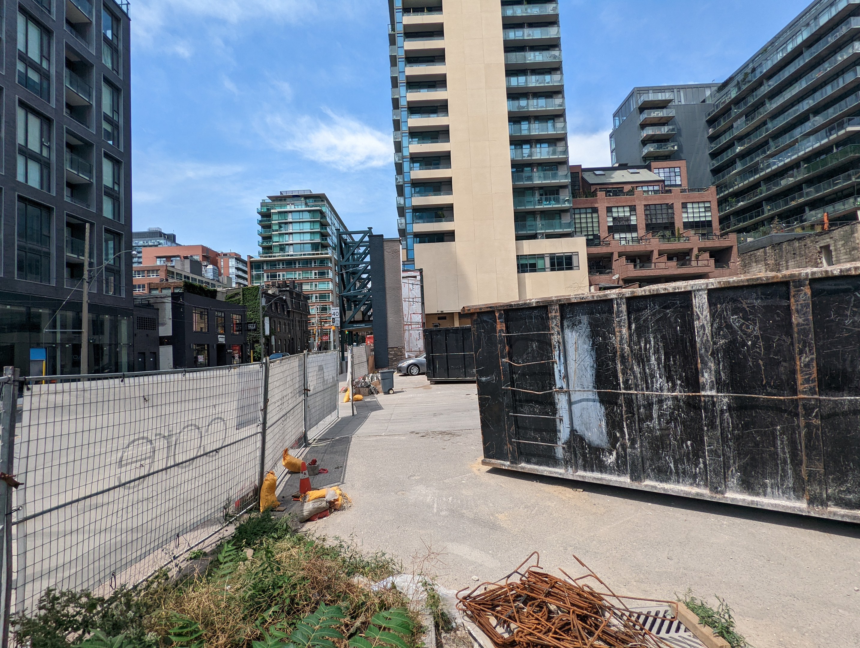 Stadtstraße mit hohen Gebäuden im Hintergrund, einem Zaun auf der linken Seite, Pflanzen unten und einem bewölkten Himmel über einer Baustelle voller Müll im Vordergrund.