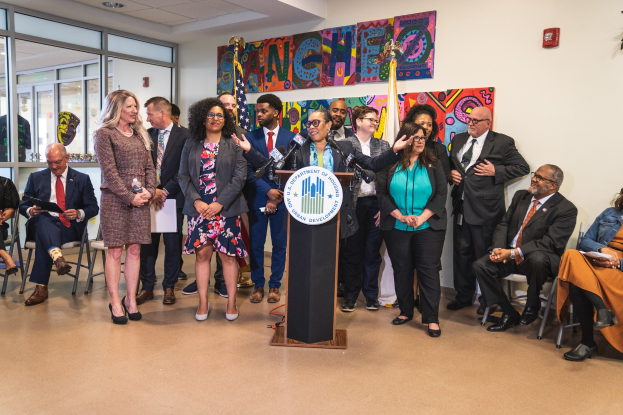 Gruppe von Menschen in einem Raum mit einem Rednerpult, einem Mikrofon und einer Tafel mit der Aufschrift "Sancho-Stadtrat tritt dem Stadtrat bei", umgeben von Stühlen, Flaggen und einem Feuerlöscher.