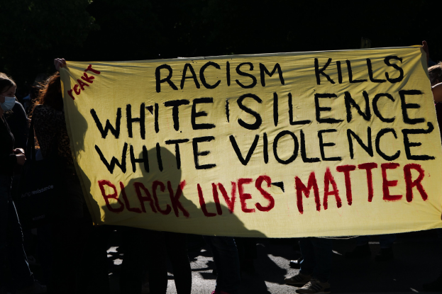 Eine Gruppe maskierter Menschen hält ein Transparent mit der Aufschrift "Rassismus tötet Weißes Schweigen, weiße Gewalt, Black Lives Matter" auf einer dunklen Straße.