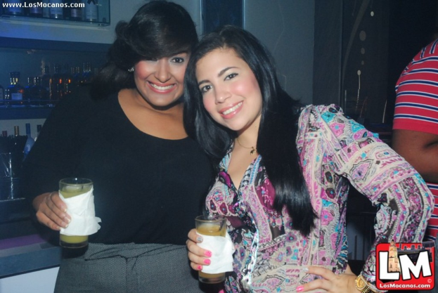 Zwei Frauen mit Gläsern und Taschentüchern, mit vielen Flaschen auf Regalen im Hintergrund und sichtbaren Wasserzeichen auf dem Bild.