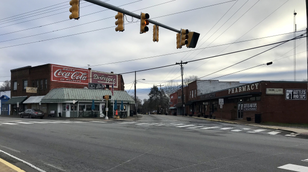 Stadtstraßenkreuzung mit einer Ampel, Fahrzeugen, Gebäuden mit Schildern, Strommasten mit Kabeln, Bäumen und einem bewölkten Himmel.