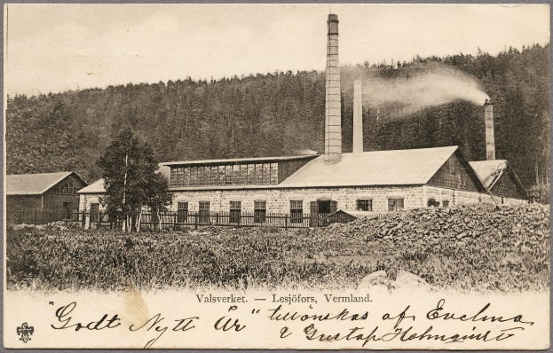 Altes Schwarz-Weiß-Foto einer Fabrik mit Rauch aus Schornsteinen, umgeben von Häusern, Bäumen, Pflanzen und einem Zaun, mit Text unten.