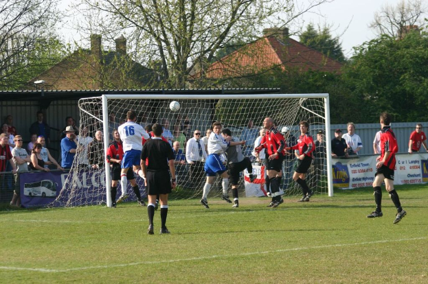 Fußballspieler spielen auf einem Feld mit einem Tor und einem Netz, während Zuschauer dahinter stehen, mit Bäumen und Häusern im Hintergrund.