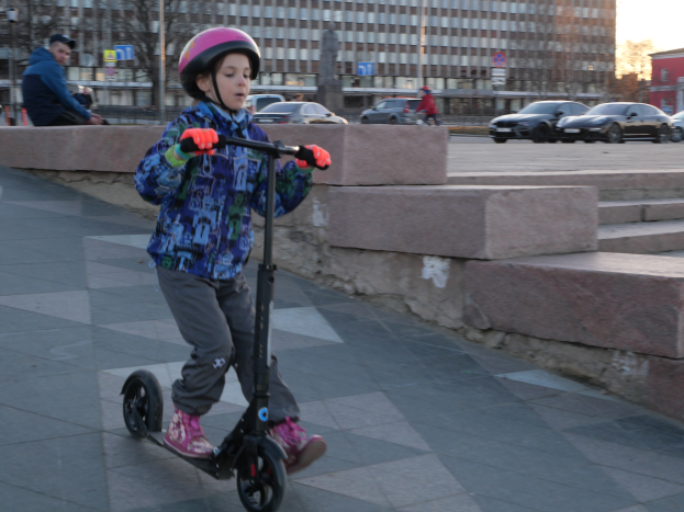 Ein junger Junge fährt mit einem Roller über einen Gehweg, trägt einen Helm und Handschuhe und hat verschiedene urbane Elemente und einen klaren blauen Himmel im Hintergrund.