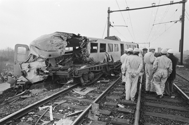 Eine Schwarz-Weiß-Szene eines Zugunfalls mit Menschen in Helmen drumherum, Strommasten, Bäumen, Gebäuden und einem klaren Himmel im Hintergrund.