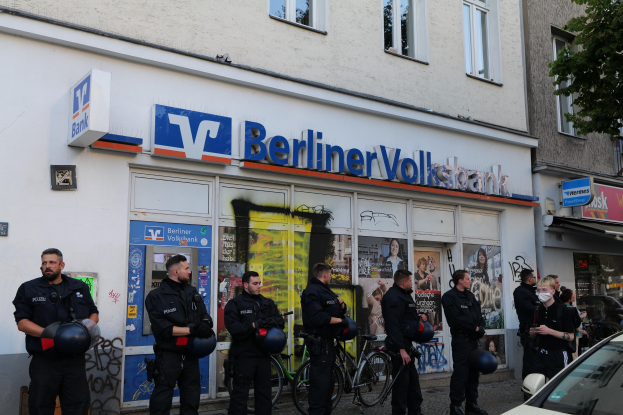 Gruppe von Polizisten vor einer Berliner Volksbank stehend, einige mit Helmen, mit einem Auto auf der rechten Seite und einem Baum auf der linken Seite, Gebäuden mit Fenstern und Texttafeln im Hintergrund.