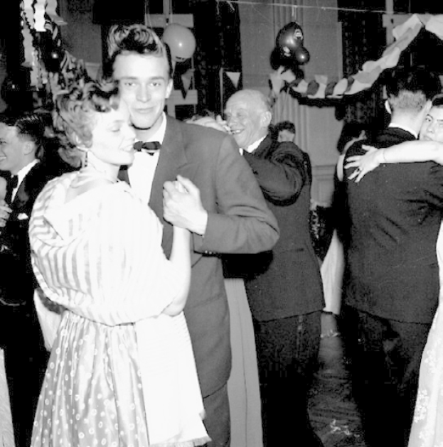 Ein Schwarz-Weiß-Foto von einem Mann und einer Frau, die auf einer Party tanzen, umgeben von lachenden Zuschauern, mit Ballons und Dekorationen im Hintergrund.