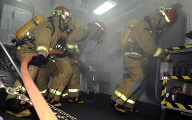 Feuerwehrleute in Schutzausrüstung, einschließlich Helmen und Sauerstoffflaschen, arbeiten an einem Feuerwehrauto mit einem Gestell von Gegenständen und Hintergrundausrüstung im Hintergrund.