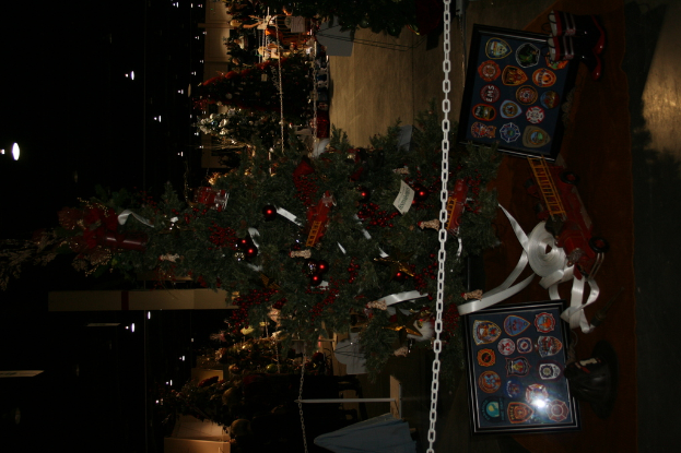 Ein Weihnachtsbaum mit Geschenken, Rahmen und Spielzeug auf einer Plattform, mit Menschen, zusätzlichen Weihnachtsbäumen und Dekorationen im Hintergrund.