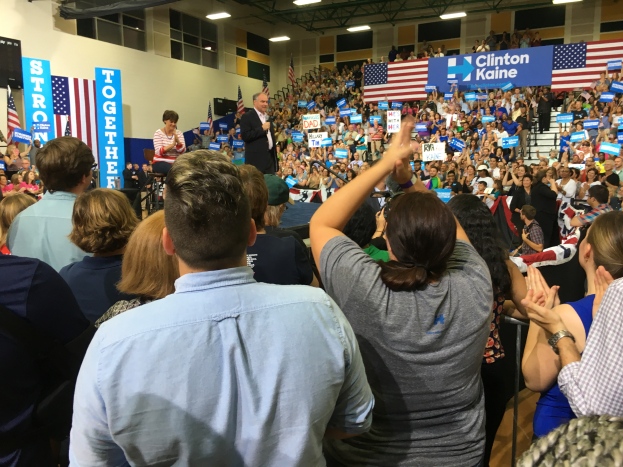Eine große Menschenmenge steht vor einer Bühne, auf der ein Mann ein Mikrofon hält und spricht, neben ihm eine Frau; die Bühne zeigt Flaggen, Banner, einen Lautsprecher, eine Wand, Fenster, Deckenleuchten und Metallstangen, während die Menge klatscht und jubelt.