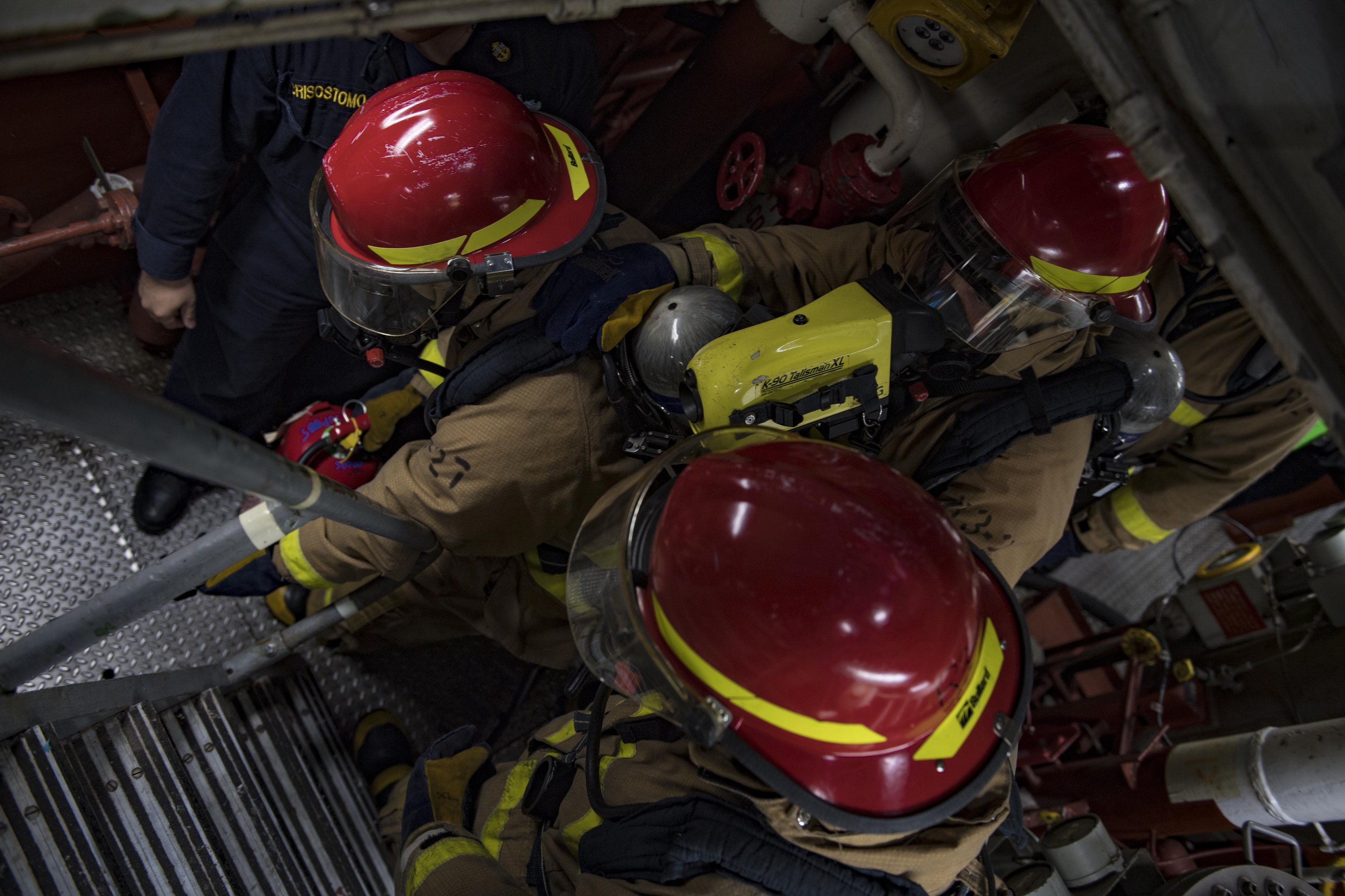 Feuerwehrleute in Helmen und Schutzausrüstung an einem Feuerwehrauto mit Schläuchen und Equipment im Hintergrund.