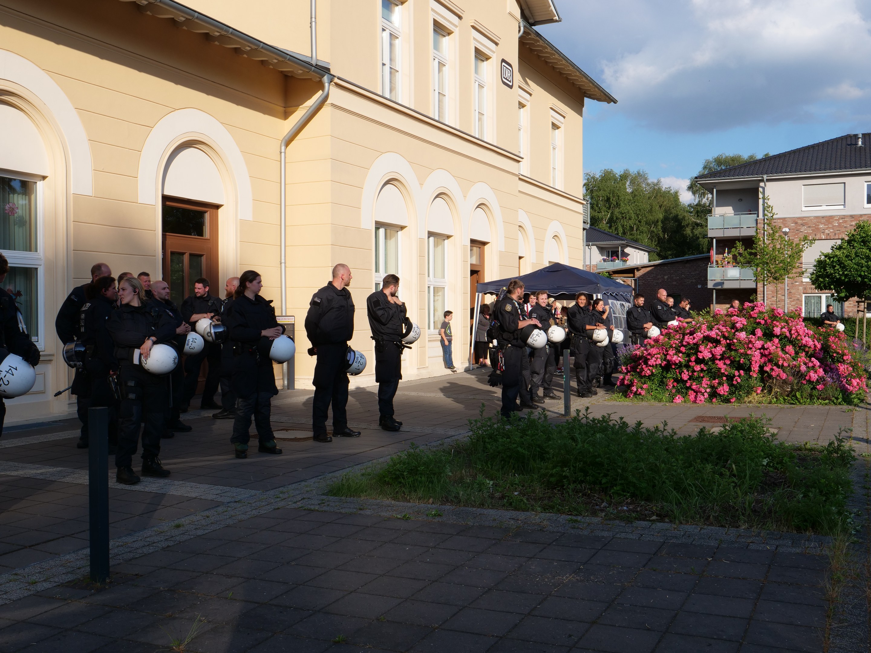 Gruppe von Polizeibeamten vor einem Gebäude mit Fenstern und Türen, Helme haltend, in der Nähe eines Zeltes, umgeben von Pflanzen, einem Straßenschild, Bäumen und einem bewölkten Himmel.