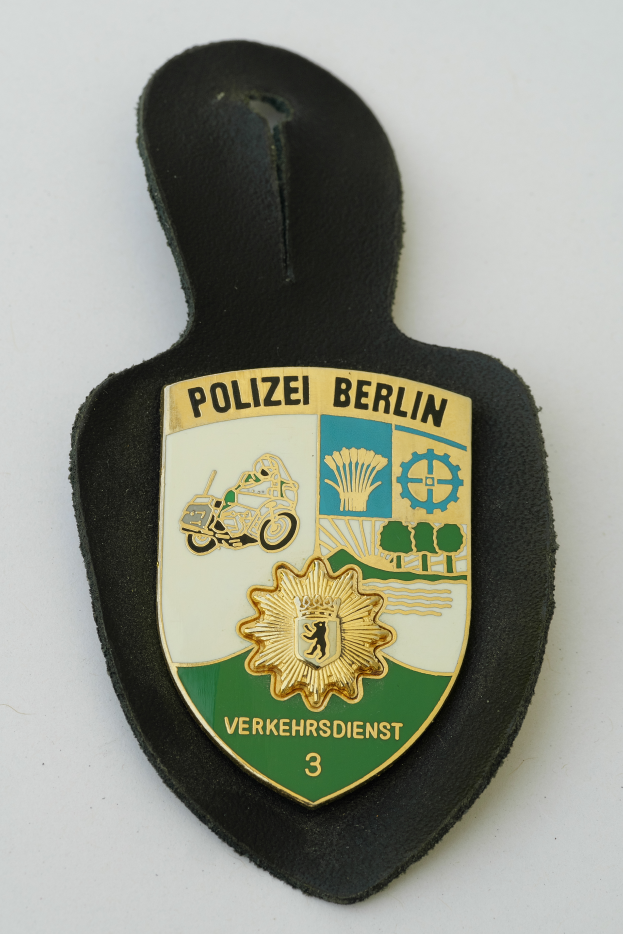 Ein Polizeidienstabzeichen mit der Aufschrift "Polizei Berlin" in fetter schwarzer Schrift auf einem blauen Schild mit einem weißen Stern und weißem Rand.