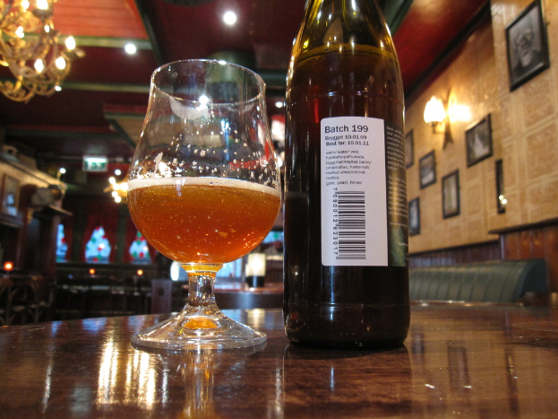 Ein Glas Wein und eine Weinflasche mit Etikett stehen auf einem Tisch, mit gerahmten Bildern, Lampen, einem Kronleuchter und einer Couch im Hintergrund.