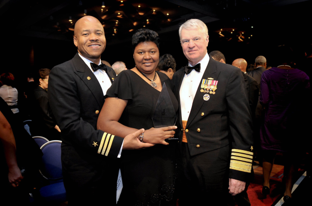 Zwei Männer in schwarzen Blazern und weißen Hemden stehen neben einer Frau in einem schwarzen Kleid, die einen Preis auf einem roten Teppich hält, während andere sitzen und stehen in einer beleuchteten Innenraumkulisse.