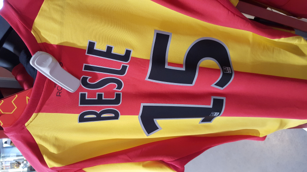 Ein roter und gelber Fußballtrikot mit Text und Zahlen darauf hängt an einem Ständer in einem Geschäft, mit anderen Gegenständen und dem Boden im Hintergrund.