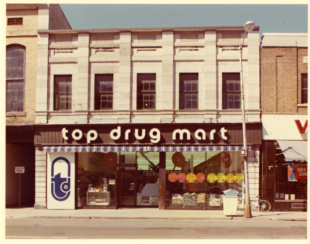 Top-Drogenladen mit Schaufenster an einer Straßenecke mit einem Schild, einem geparkten Fahrrad, einem Müllcontainer, einem Straßenpfahl und einem bewölkten Himmel.