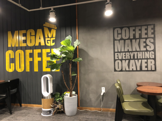 Ein Kaffeehaus-Interieur mit Tischen, Stühlen, einem "Mega Coffee"-Schild an der Wand, einer Bodenstehmaschine, Deckenleuchten und einem Topfgewächs.