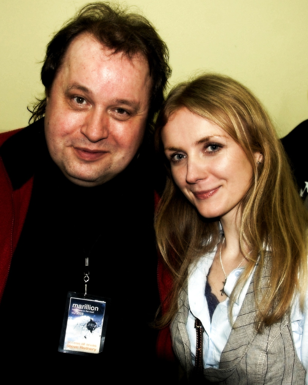 Ein Mann und eine Frau lächeln, mit einem Abzeichen sichtbar, vor einem hellgelben Hintergrund.