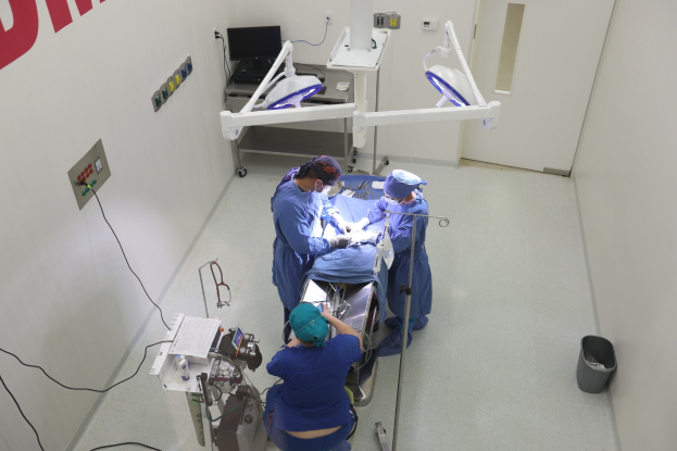 Eine Gruppe von Chirurgen führt einen Eingriff in einem Operationssaal durch, wobei ein Patient auf einer Trage von medizinischer Ausrüstung umgeben ist, eine textbedeckte Wand auf der linken Seite, ein Objekt auf dem Boden auf der rechten Seite und verschiedene medizinische Geräte im Hintergrund.