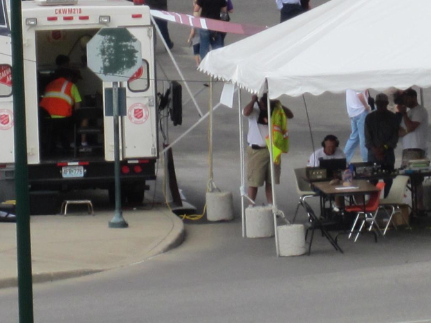 Ein Krankenwagen steht links von einer StraÃŸe, wÃ¤hrend auf der rechten Seite ein Zelt viele Menschen schÃ¼tzt, mit einem Tisch und einem Stuhl in der NÃ¤he.