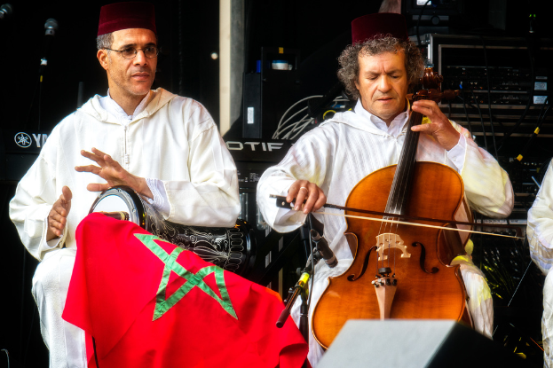 Zwei Männer in passenden Kostümen und Mützen sitzen und spielen verschiedene Musikinstrumente, mit sichtbaren Musikanlagen dahinter.