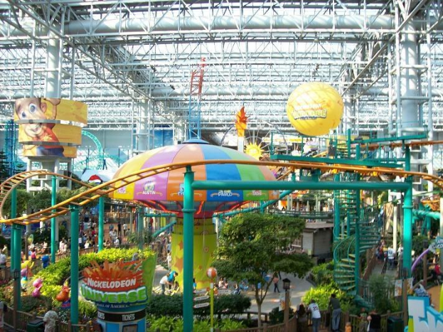 Indoor-Wasserpark mit einer Achterbahn, umgeben von Menschen, Bäumen, Pflanzen und Deckenleuchten mit Stangen.
