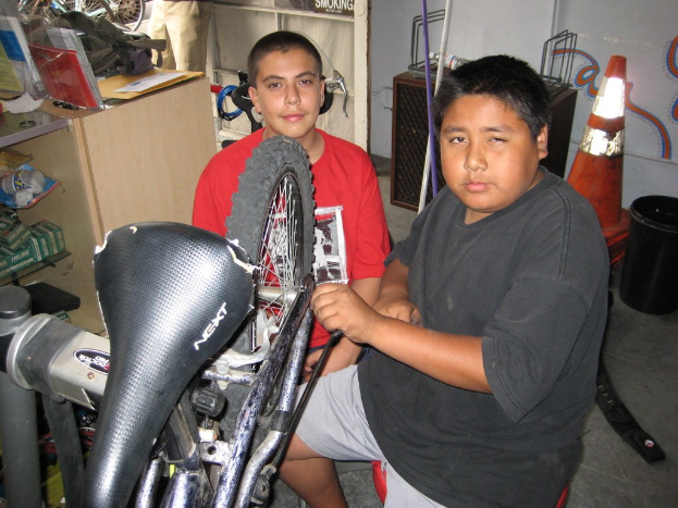 Zwei Personen reparieren ein Fahrrad in der Mitte, während Büromaterial und Fahrräder im Hintergrund zu sehen sind.