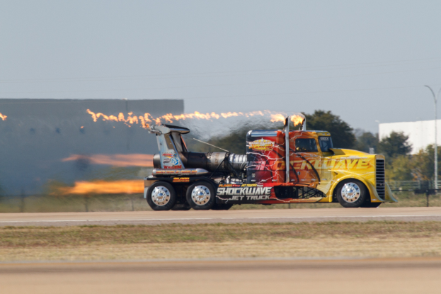 Ein Lkw mit Flammen, die aus seinem Heck austreten, f├Ąhrt auf einer von Gras, einem Zaun, B├Ąumen, einem Geb├Ąude, einem ├ťberlandungsmast, Dr├Ąhten und einer bew├Âlktem Himmel ges├Ąumten Stra├če, mit einem sichtbaren Reifen und Rad.