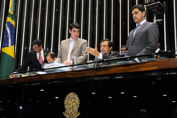 Leute, die an einem Tisch auf einer Bühne mit einer Holzpaneelwand und Fahnen dahinter sitzen und ein goldenes Logo auf dem Tisch vorne.
