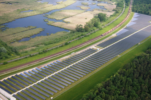 Luftaufnahme einer Solar-Farm mit Panelen, umgeben von Bäumen, Gras, Pflanzen und Wasser, mit einem Zug auf einer nahen Bahnlinie.