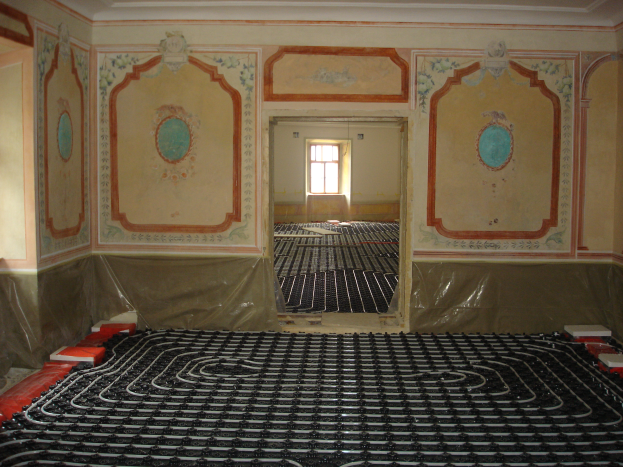 Ein Raum mit einer Fußbodenheizung in der Mitte, umgeben von Wänden und einem Fenster im Hintergrund, mit verschiedenen Gegenständen auf dem Boden, die wahrscheinlich mit dem Heizungssystem zu tun haben.