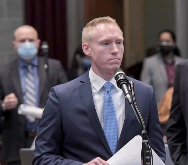 Ein Mann im Anzug spricht an einem Podium mit Papieren vor einem Mikrofon, während andere in der Nähe gegen eine Vorhangwand stehen.