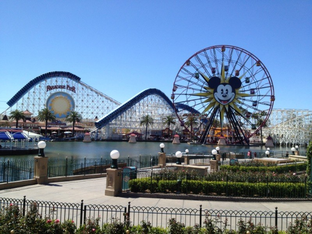 Ein Blick auf Disneylands California Adventure mit einem Riesenrad, einer Achterbahn, Grünflächen, Laternenmasten, einem Gewässer und einem hellblauen Himmel.