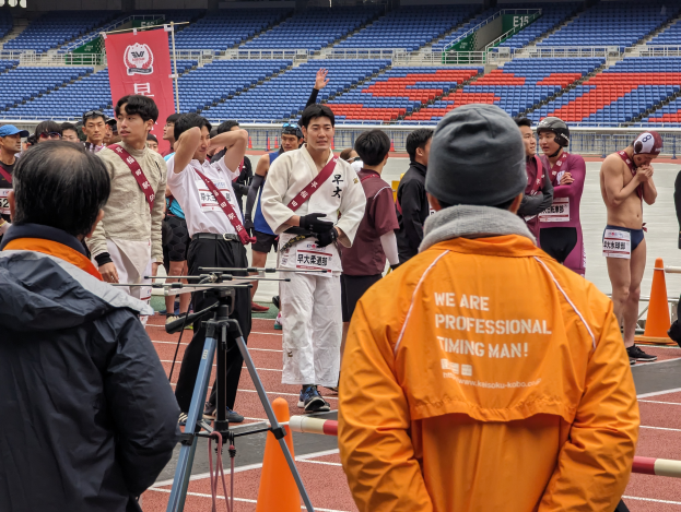 Eine Gruppe von Menschen, die auf einer Bahn stehen, mit einer Kamera auf Stativ im Vordergrund, Stühlen, einem Banner und Lampen im Hintergrund.