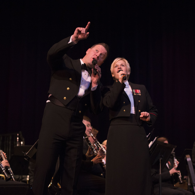 Ein Mann und eine Frau in Fracks, die Mikrofone halten, auf einer Bühne mit einem sitzenden Orchester im Hintergrund vor einem dunklen Hintergrund.