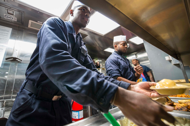 Eine Gruppe von Männern steht in einer Küche, einer hält einen Teller mit Essen, mit Tabletts voller Essen auf dem Tisch, einem Feuerlöscher und Papieren an der Wand im Hintergrund.