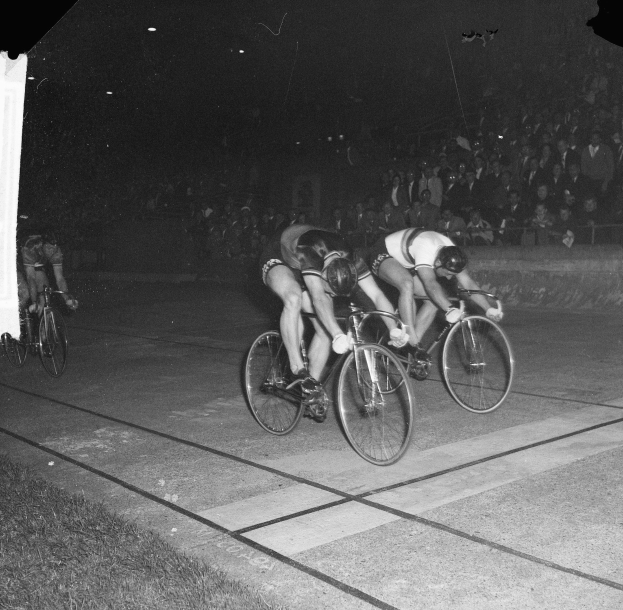 Schwarzes und weißes Bild einer Gruppe von Männern, die nachts auf Fahrrädern eine Straße entlangfahren, mit einer Menge von Menschen im Hintergrund und von Straßenlaternen beleuchtet.