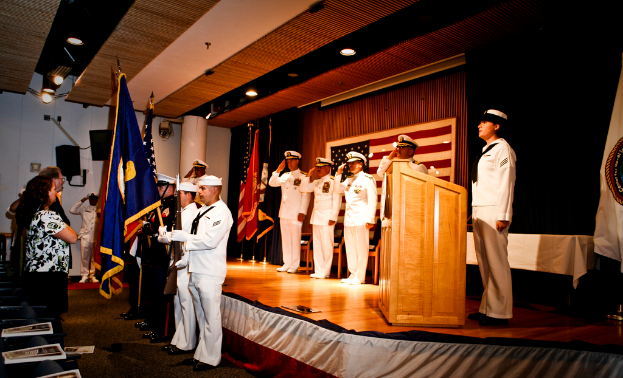 Eine offizielle Veranstaltung mit Menschen auf einer Bühne, die salutieren, sowie Flaggen in Rot, Blau und Weiß, Stühle, ein Rednerpult und ein Tisch mit einer weißen Decke, sowie im Hintergrund ein Fernseher und eine Lautsprecherbox.