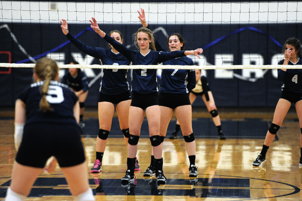 Drei Mädchen bei einem Volleyballspiel auf dem Feld stehend mit erhobenen Händen, ein Netz davor und ein Zaun im Hintergrund.